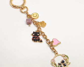Puede incluir: Llavero dorado con múltiples colgantes. Los colgantes incluyen un gato negro con corazones rojos, un corazón rosa, una huella de pata dorada, una cuenta morada y un colgante circular. El llavero también presenta una corona con dos gatos negros y flores rosas.