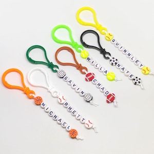 May include: A selection of personalised sports-themed keyrings. Each keyring has a different coloured clip and a string of white beads spelling out a name, with sports ball charms such as basketballs, baseballs, and footballs.