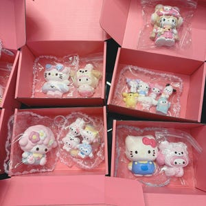 May include: A collection of Hello Kitty figurines in pink boxes. The figurines are in various poses and outfits, including dresses and overalls. Each figurine is individually packaged in a clear plastic bag with a lace trim.