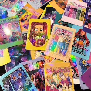 May include: A collection of colorful trading cards featuring anime-style illustrations. The cards display various characters and the text "SAJA BOYS", "HUNTRIX", and "MIRA". Some cards have a holographic finish, adding a shimmering effect.