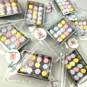 Könnte beinhalten: Mehrere transparente Plastiktüten, die jeweils ein kleines silbernes Backblech mit pastellfarbenen, pfotenförmigen Süßigkeiten enthalten. An jeder Tüte ist ein kleiner rosa Bärenanhänger mit dem Text "Waku Squishy" befestigt.