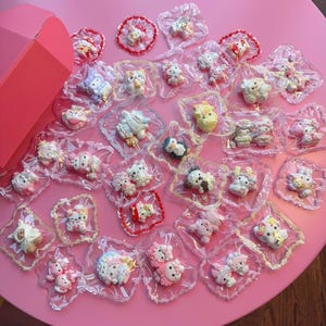 May include: A selection of small, colourful character-shaped decorations, individually packaged in clear plastic. The characters, including animals and other cute figures, are arranged on a pink surface with a pink box in the background.