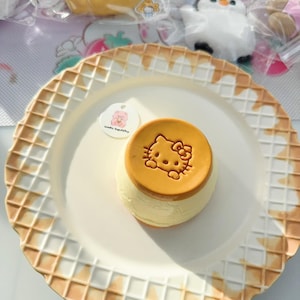 May include: A white ceramic plate with a waffle pattern border. The plate has a small, round, white sticker with the text "Cutie Tasty" and a Hello Kitty-themed pudding with a brown caramel topping.