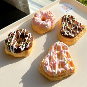 May include: Four decorated donuts on a white plate. The donuts are in the shapes of a heart, a circle, and two irregular shapes. The donuts are decorated with pink, white, and brown frosting and colorful sprinkles.