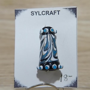 May include: A black and white glass bead with a swirl pattern and blue accents. The bead is on a white card with the word "SYLCRAFT" printed on it.