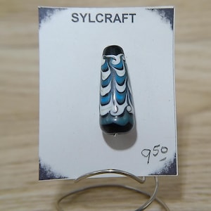 May include: A blue and white glass bead with a swirl pattern. The bead is on a white card with the text "SYLCRAFT" and "950" written on it.