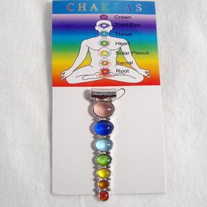 May include: A silver charm with seven colored stones representing the seven chakras. The stones are pink, blue, light blue, green, yellow, orange, and red. The charm is on a white card with the word "CHAKRAS" and a diagram of a person with the chakras labeled.