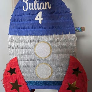 Könnte beinhalten: Eine blaue und silberne raketenförmige Piñata mit roten Flügeln, die mit goldenen Sternen verziert sind. Die Piñata ist mit "Julian 4" beschriftet.