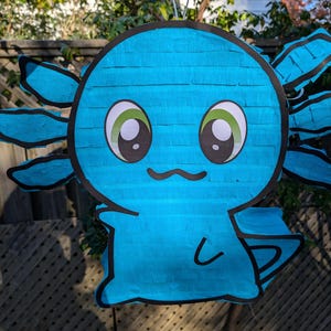 May include: A blue axolotl piñata with black outlines and a smiling face. The piñata has large, cartoonish eyes with green and white details. It is suspended by a metal wire, ready for a party or celebration.