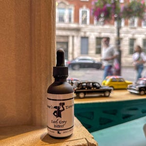 May include: A small black glass bottle with a dropper top. The bottle is labeled "Tavem's Earl Grey Bitters" and has a small illustration of a person holding a hammer. The bottle is sitting on a brick surface.