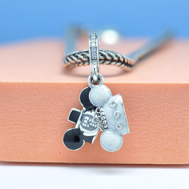May include: A silver charm with a Mickey Mouse design. The charm features black and white enamel details, with a small cluster of clear stones. The charm is suspended from a silver chain.
