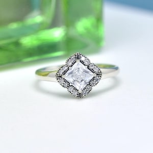May include: A silver ring with a square, clear gemstone at the center, surrounded by smaller, round, clear stones. The setting is a four-leaf clover shape. The ring is on a white surface with a blurred green background.