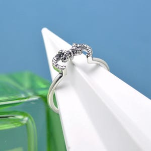 May include: A silver ring featuring a bow design, embellished with tiny, glittering stones. The ring is presented on a white surface, set against a blue backdrop, which accentuates the ring's intricate details.