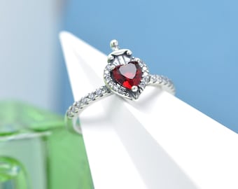 May include: A silver ring with a heart-shaped, deep red gemstone at the center, surrounded by smaller clear stones. The ring has a crown-like detail above the heart and is set on a white surface against a blue background. A piece of jewelry.