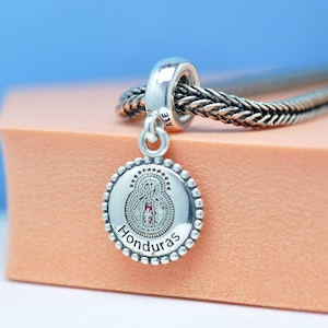Peut inclure: Un charm en argent avec un pendentif rond représentant une image d'une figure religieuse et le mot "Honduras". Le charm est attaché à une chaîne en argent. Le pendentif a une bordure perlée et un design détaillé.