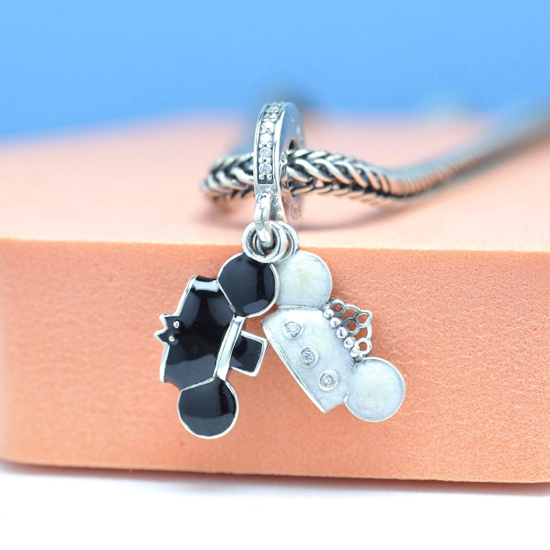May include: A silver charm with a black and white tuxedo and wedding dress design. The charm features a small loop with clear stones and is attached to a silver chain. The charm is likely a wedding or anniversary gift.
