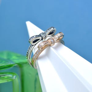 May include: Two rings with bow designs, one silver and the other gold, are displayed. Both rings feature small, clear gemstones set along the band and bow. The rings are positioned on a white, angular surface against a blue background.