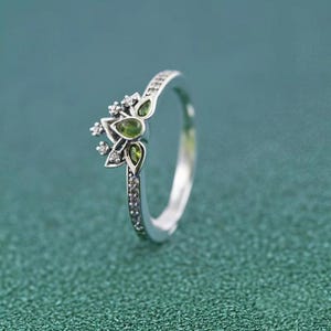 May include: A silver ring with a floral design, featuring green and clear gemstones. The ring's band is embellished with small, clear stones. The main feature is a cluster of green, leaf-shaped stones and clear, round stones.