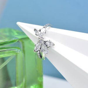 Puede incluir: Un anillo plateado con un diseño floral, con múltiples piedras preciosas claras y brillantes. El anillo se presenta sobre una forma geométrica blanca con un fondo azul claro, mostrando los intrincados detalles de la joya.