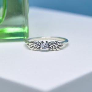 Puede incluir: Un anillo de plata con un dise&ntilde;o de alas de &aacute;ngel. El anillo presenta una piedra preciosa central transparente y piedras m&aacute;s peque&ntilde;as a lo largo de las alas. El anillo est&aacute; sobre una superficie blanca con un fondo verde y azul.