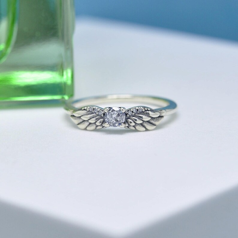 Puede incluir: Un anillo de plata con un dise&ntilde;o de alas de &aacute;ngel. El anillo presenta una piedra preciosa central transparente y piedras m&aacute;s peque&ntilde;as a lo largo de las alas. El anillo est&aacute; sobre una superficie blanca con un fondo verde y azul.