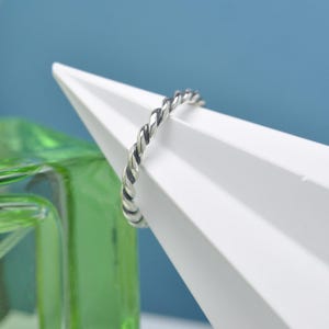 May include: A silver-coloured, twisted rope ring. The ring is set against a white geometric shape and a green glass object, with a blue background. The ring's design features a braided or twisted pattern, creating a textured appearance.