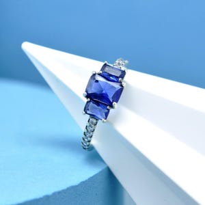 Peut inclure: Une bague en argent ornée de trois pierres précieuses rectangulaires bleu foncé. La monture est ornée de petites pierres claires. La bague est présentée sur une forme géométrique blanche et un fond bleu.