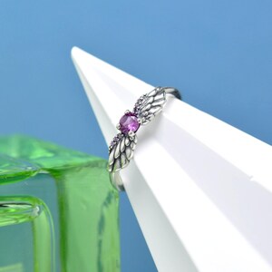 Puede incluir: Un anillo plateado con un dise&ntilde;o de alas de &aacute;ngel, con una piedra preciosa central redonda y p&uacute;rpura y piedras de acento p&uacute;rpura m&aacute;s peque&ntilde;as. El anillo est&aacute; colocado contra una forma geom&eacute;trica blanca y un fondo azul.