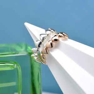 May include: Two heart-shaped rings, one silver and one rose gold, are displayed on a white geometric stand against a blue background. The rings are composed of small, connected hearts, showcasing a romantic and delicate design. The rings are a jewelry item.