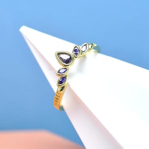 May include: A gold-colored ring with a leaf-like design. The ring features a large, teardrop-shaped purple gemstone at the center, surrounded by smaller, oval-shaped purple gemstones. The band has a textured pattern.