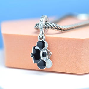 May include: A silver charm with a black and white design, featuring a stylized shape with a small star and a round element. The charm is suspended from a silver chain and has a small loop with clear stones.