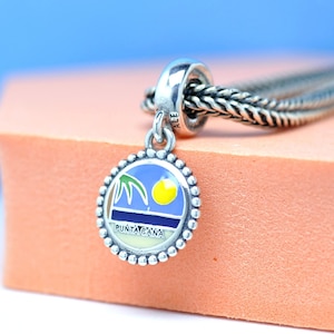 Op de afbeelding: Een zilveren bedel met een ronde hanger met een strandscène met een palmboom, zon en de tekst "PUNTA CANA". De bedel is bevestigd aan een gevlochten zilveren ketting. De hanger heeft een kralenrand.