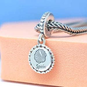 May include: A silver charm featuring a seashell design and the word "Spain". The charm is attached to a silver bracelet. The background is a light pink and blue.