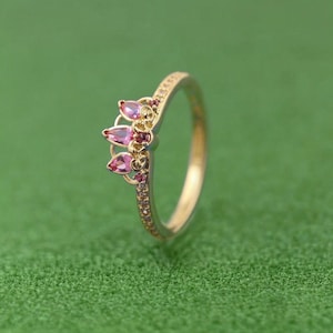 Può includere: Un anello color oro con un design a corona con tre gemme rosa a forma di goccia. La fascia dell'anello è impreziosita da piccole pietre rosa rotonde. L'anello è posto su uno sfondo verde.