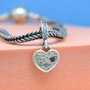 May include: Silver charm with a heart-shaped pendant reading "But First, Coffee." The charm features a coffee cup design and is attached to a silver bracelet. The charm is embellished with small, clear stones.
