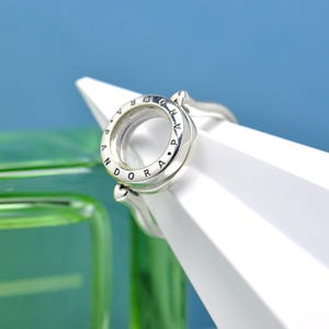 Puede incluir: Un anillo plateado con la palabra "Pandora" grabada alrededor del borde exterior. El anillo tiene un diseño circular con un centro transparente y está colocado sobre una forma geométrica blanca y un fondo azul.