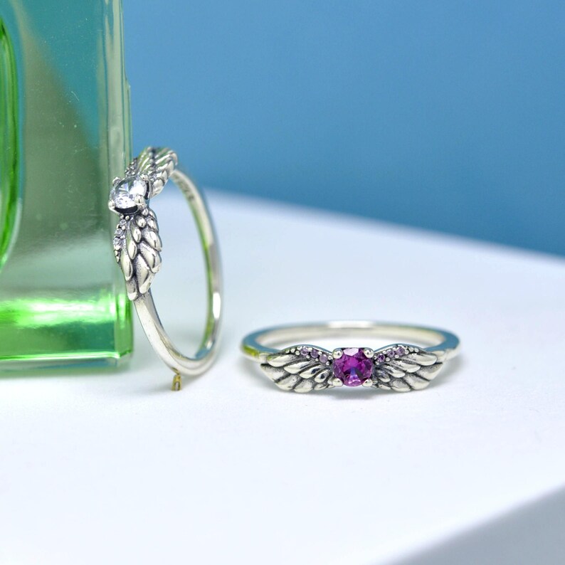 Puede incluir: Dos anillos de plata con dise&ntilde;os de alas. Un anillo presenta una piedra preciosa transparente, mientras que el otro tiene una piedra preciosa p&uacute;rpura en el centro. Ambos anillos tienen peque&ntilde;as piedras de acento transparentes. Los anillos se exhiben sobre una superficie blanca con un fondo azul.