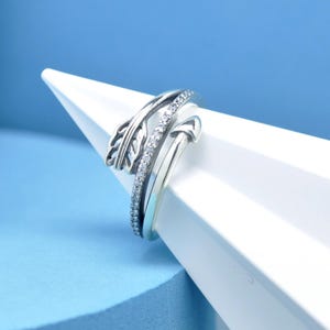 May include: A silver ring with a feather design and a band adorned with small, clear stones. The ring is displayed on a white, geometric shape against a blue background. The ring's design includes an arrow-like shape.