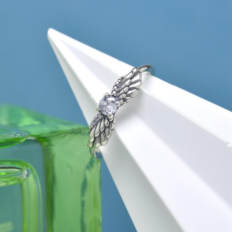 Puede incluir: Un anillo plateado con un dise&ntilde;o de alas de &aacute;ngel. El anillo tiene una piedra preciosa central transparente y piedras de acento m&aacute;s peque&ntilde;as en las alas. Las alas est&aacute;n detalladas con texturas similares a plumas. El anillo se muestra sobre un fondo blanco y azul.