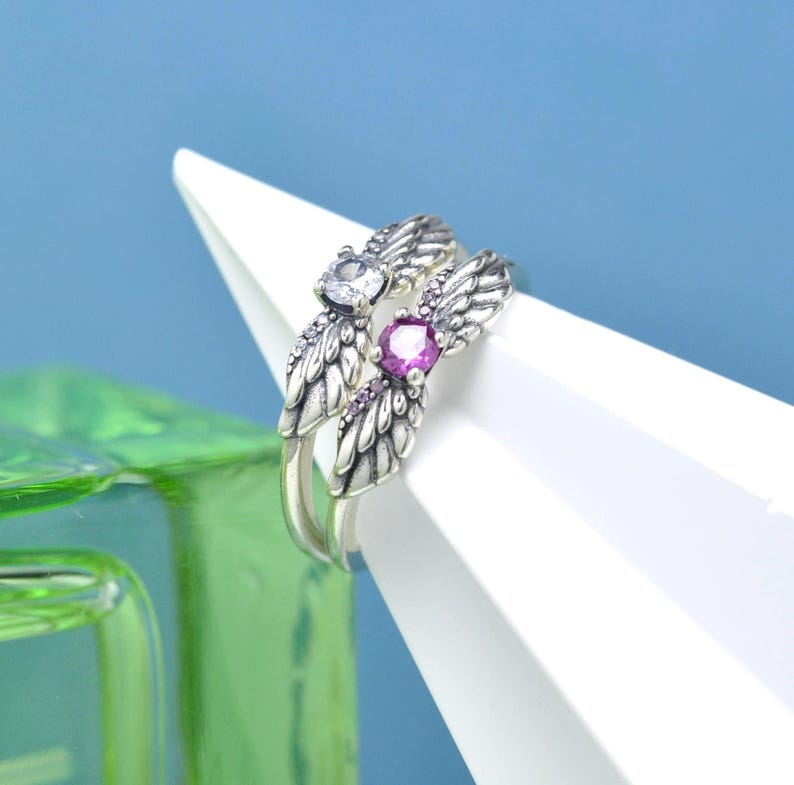 Puede incluir: Un anillo plateado con dos dise&ntilde;os de alas de &aacute;ngel, cada uno adornado con peque&ntilde;as piedras transparentes. Un ala presenta una gran gema redonda de color p&uacute;rpura. El anillo se muestra sobre una superficie blanca angular sobre un fondo azul.