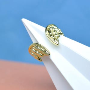 May include: A pair of gold-colored wing-shaped earrings. The earrings are positioned on a white surface against a blue and pink background. The wings have detailed feather patterns.