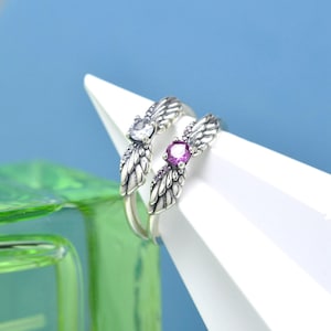 Op de afbeelding: Twee zilveren ringen met vleugelontwerpen. Eén ring heeft een heldere edelsteen en de andere een paarse edelsteen. De ringen worden getoond op een witte geometrische vorm tegen een blauwe achtergrond.