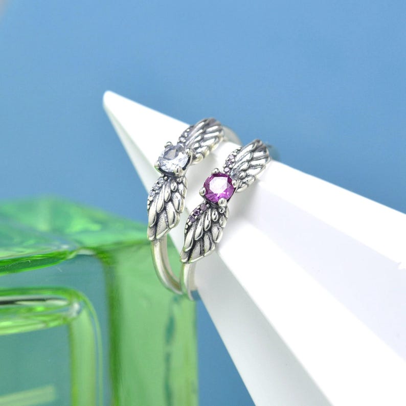 Puede incluir: Dos anillos de plata con dise&ntilde;os de alas. Un anillo presenta una gema transparente y el otro una gema p&uacute;rpura. Los anillos se exhiben sobre una forma geom&eacute;trica blanca sobre un fondo azul.