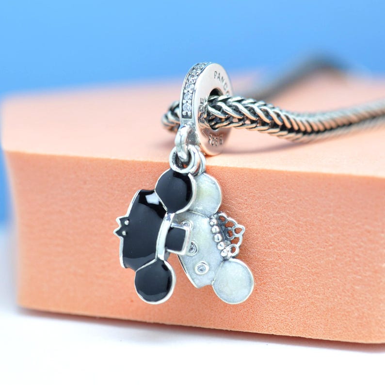 May include: A silver charm with a black and white Mickey Mouse design. The charm has a loop with clear stones and is attached to a silver chain. The charm is on a pink surface with a blue background.