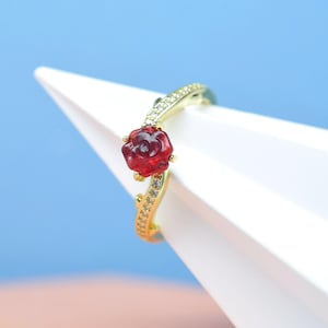 May include: A gold ring with a red rose-shaped gemstone. The band is embellished with small, clear gemstones. The ring is set against a white and blue backdrop, accentuating its elegant design.