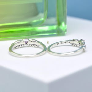 Puede incluir: Dos anillos de plata con dise&ntilde;os de plumas y peque&ntilde;as piedras preciosas. Un anillo tiene una piedra preciosa p&uacute;rpura, el otro una piedra preciosa transparente. Los anillos est&aacute;n marcados con "S925 ALE 54".