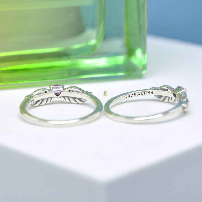 Puede incluir: Dos anillos de plata con dise&ntilde;os de plumas y peque&ntilde;as piedras preciosas. Un anillo tiene una piedra preciosa p&uacute;rpura, el otro una piedra preciosa transparente. Los anillos est&aacute;n marcados con "S925 ALE 54".