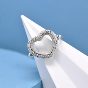 Op de afbeelding: Een zilveren ring met een hartvormig ontwerp. Het hart is omrand met kleine, heldere edelstenen. De ring wordt gepresenteerd op een wit, hoekig oppervlak tegen een blauwe achtergrond. De ring is een sieraad.