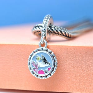 Puede incluir: Charm de plata con un colgante redondo con un diseño de criatura marina. El colgante tiene un fondo azul claro con una manta raya en blanco y negro, coral rosa y un borde festoneado.