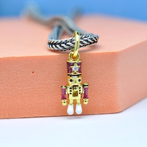 May include: A gold-colored charm pendant in the shape of a nutcracker soldier. The pendant has red, white, and gold accents, with a star-shaped design on the hat. The charm is attached to a silver-colored braided chain.